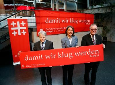 drei Funktionäre halten ein Banner mit dem Kirchentagsmotto 2015 'Damit wir klug werden'