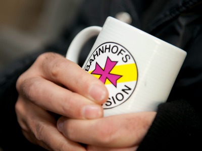 Hände halten einen Tee-Becher mit dem Logo der Bahnhofsmission
