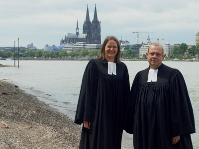 Ein Pfarrer und eine Pfarrerin im Talar am Rheinufer, im Hintergrund der Kölner Dom